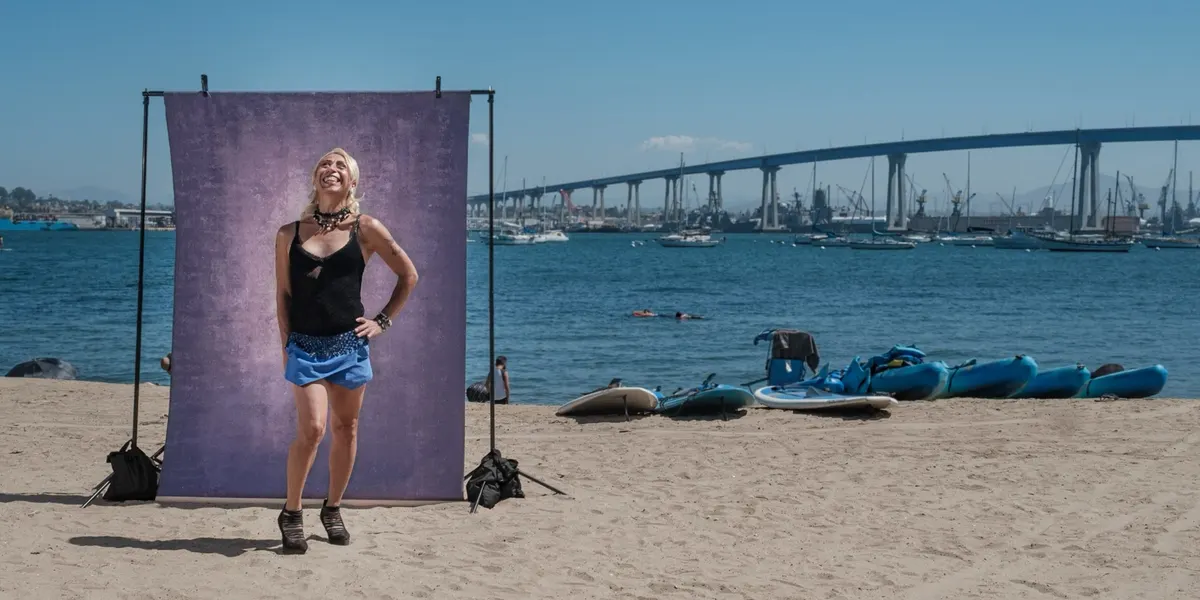 Alexis laughs with her face turned to the sky, in a black top and blue skirt with a hand on her hip, posed at a purple backdrop on the sand with the Coronado Bridge behind her.