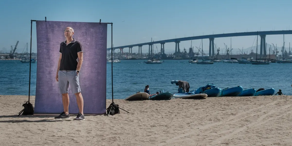 Craig gazes into the distance in a dark polo, standing before a purple backdrop on the beach with sailboats and the Coronado Bridge spanning the bay behind him.