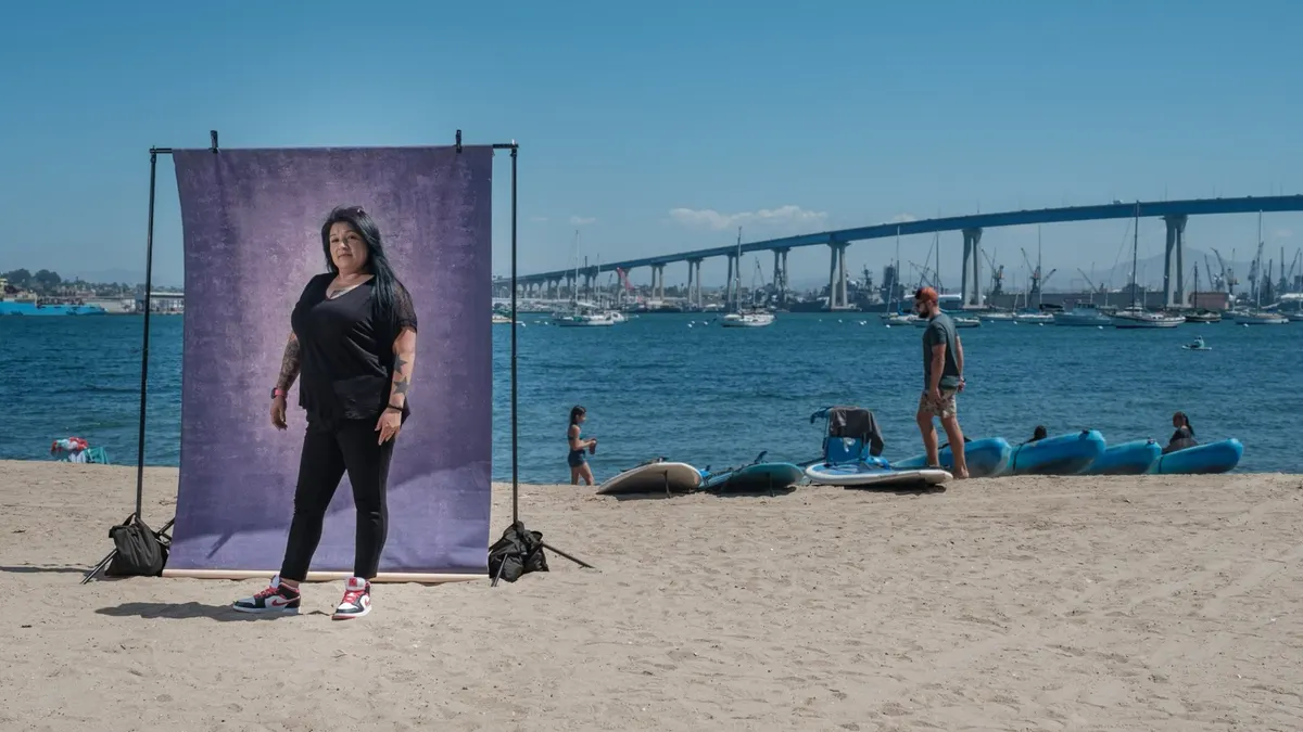 Elsa stands confidently in black with red-and-white sneakers, tattoos visible on her arms, in front of a purple backdrop on the sand with the Coronado Bridge behind her.