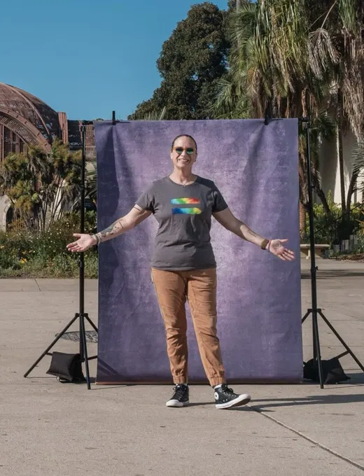 Mollie throws her arms open wide in a tee with a rainbow graphic, grinning in front of a purple backdrop at Balboa Park.