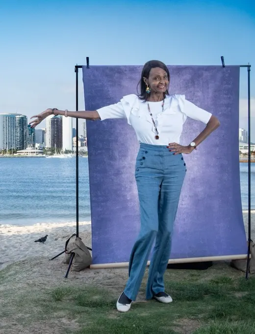Tracie strikes a playful pose in a white puff-sleeve top, one arm draped along a purple backdrop, with San Diego Bay behind her.