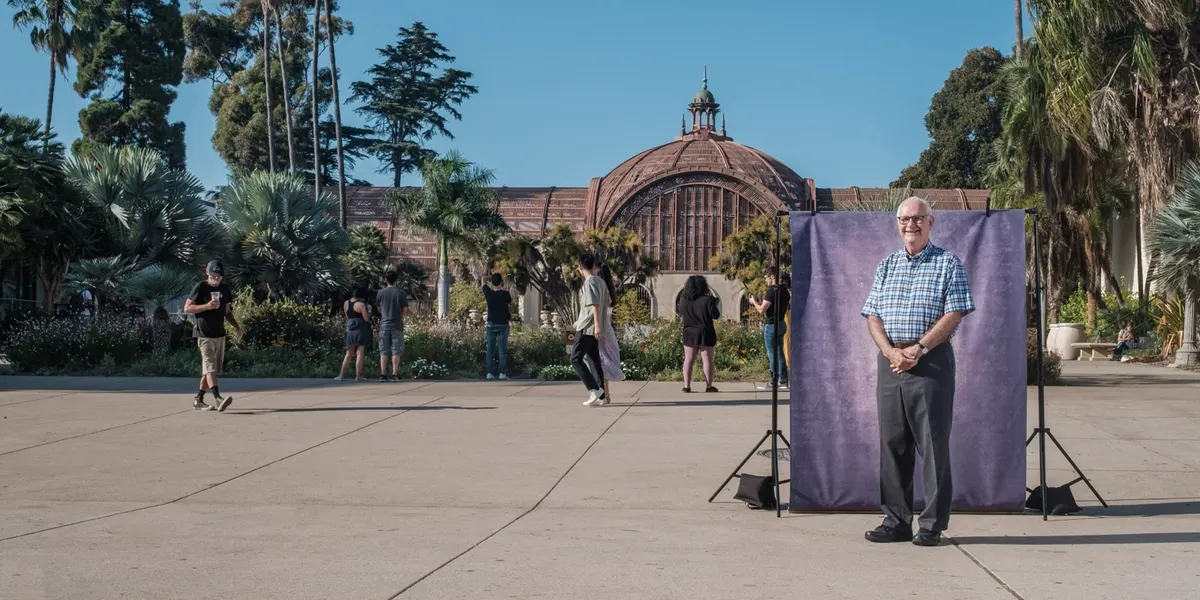 Jim smiles gently with hands clasped in front of him, dressed in a blue plaid shirt, posed at a purple backdrop on the busy Balboa Park promenade.