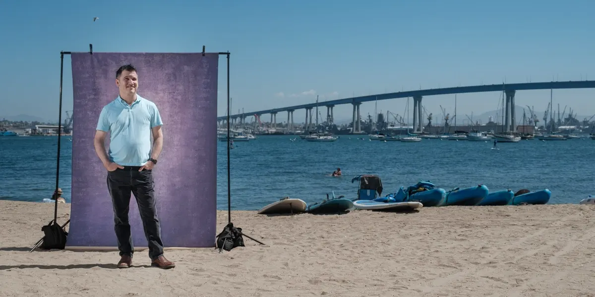 Joe smiles off to the side with hands in his pockets, wearing a light blue polo, standing at a purple backdrop on the beach with sailboats on the bay behind him.