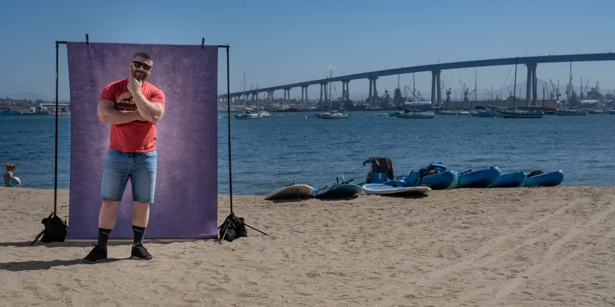 John strikes a thoughtful chin-on-fist pose in sunglasses and a red graphic tee, in front of a purple backdrop on the sand with the Coronado Bridge behind him.