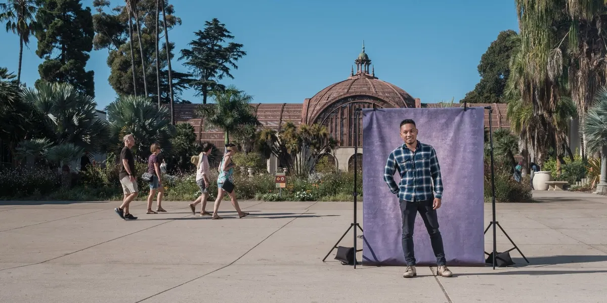 Jorome smiles with a hand on his hip in a blue plaid flannel, posed at a purple backdrop on the Balboa Park plaza near the Botanical Building.