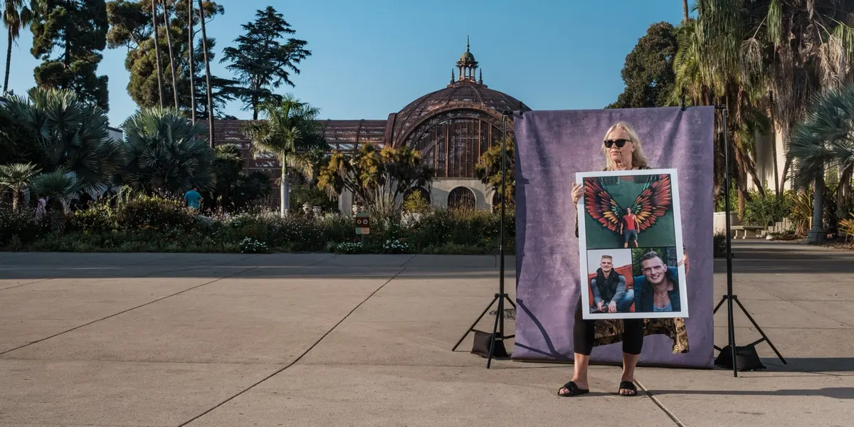 Judi holds a large framed collage of photographs close to her chest, standing quietly in front of a purple backdrop on the Balboa Park promenade.