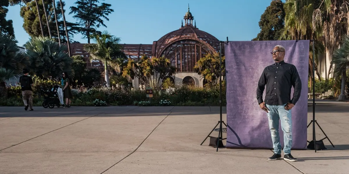 Kevin stands tall in a black button-up shirt and sunglasses, in front of a purple backdrop on the Balboa Park promenade with the Botanical Building dome behind him.