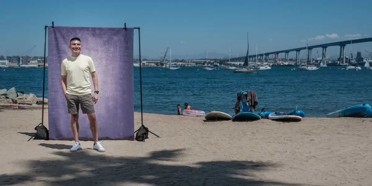 Martin smiles in a pale yellow tee and white sneakers, hands relaxed, standing at a purple backdrop on the beach with the Coronado Bridge stretching across the water.