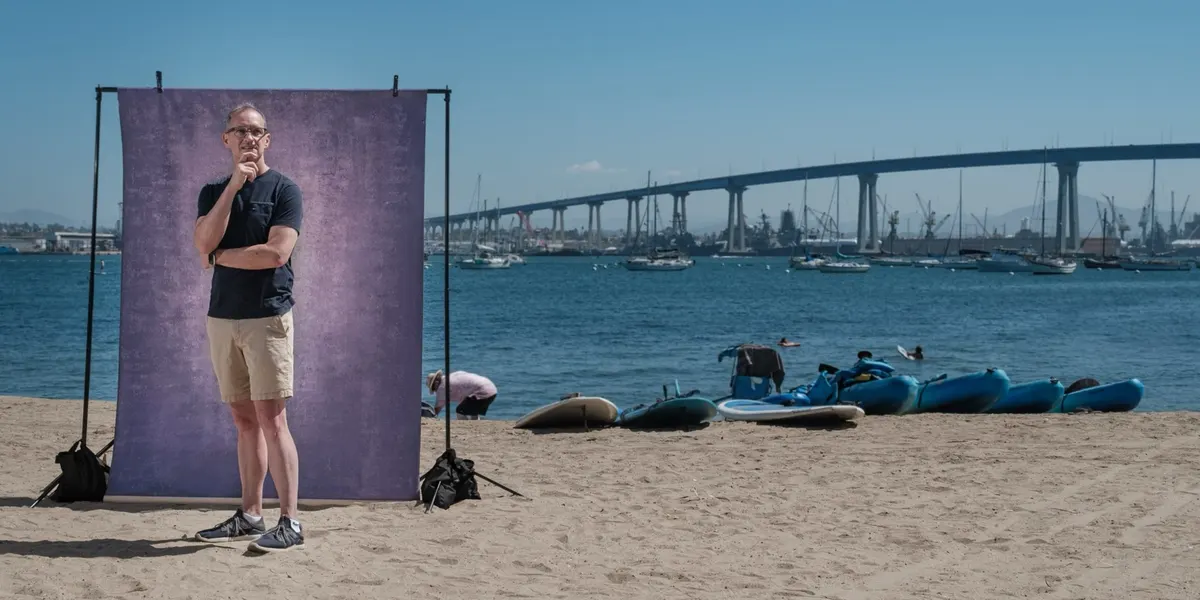 Michael poses chin in hand in a navy tee, standing at a purple backdrop on the sand with sailboats and the Coronado Bridge filling the bay behind him.