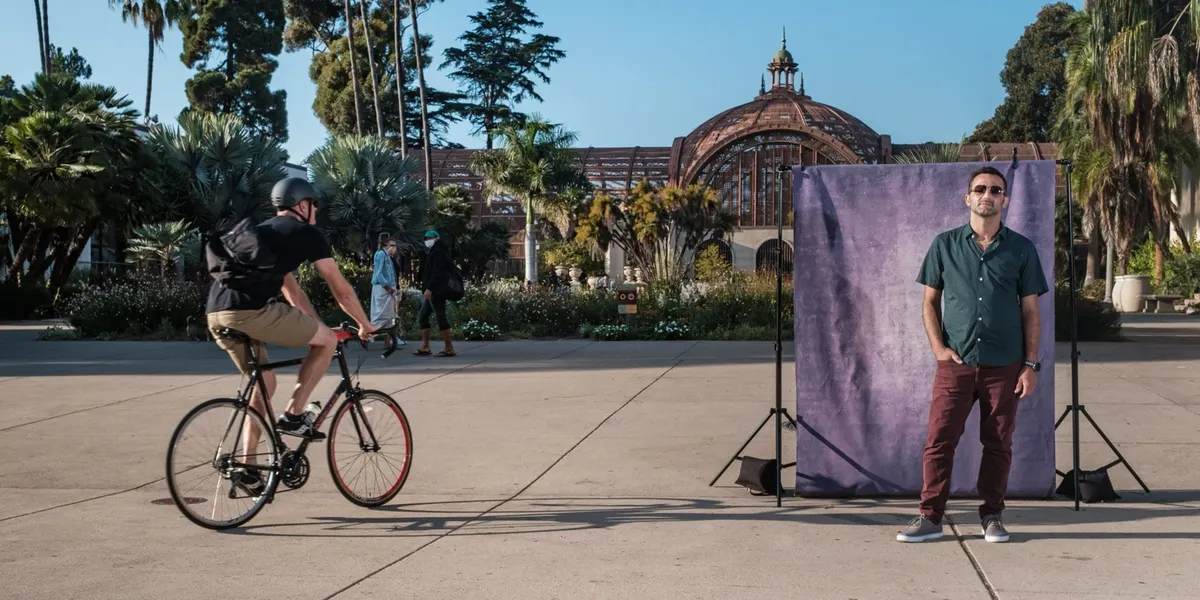 Roger stands cool in sunglasses, a dark green shirt, and rust-colored pants at a purple backdrop on the Balboa Park plaza, a cyclist rolling past beside him.