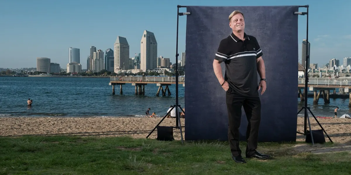 Shawn beams with his hands in his pockets, wearing a striped polo, in front of a navy backdrop on the Coronado waterfront with the San Diego skyline in the distance.