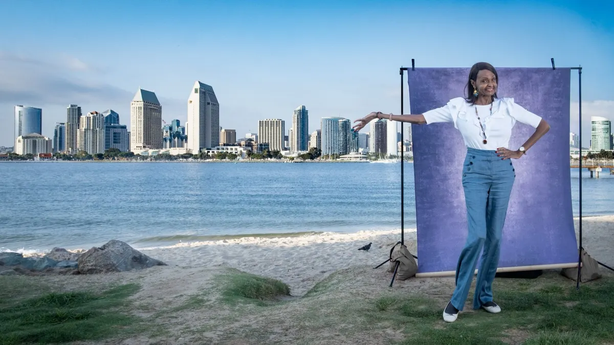 Tracie strikes a playful pose in a white puff-sleeve top, one arm draped along the top of a purple backdrop, with San Diego Bay and the downtown skyline behind her.